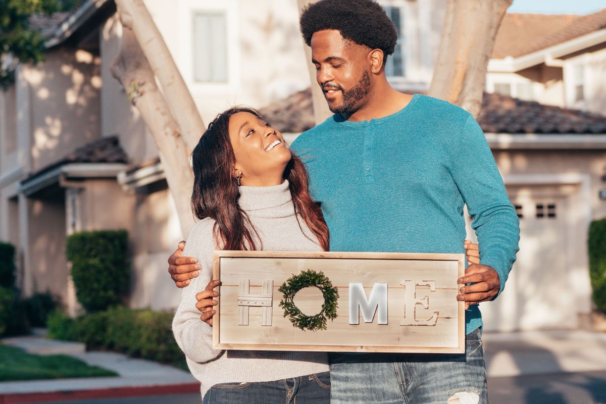 Couple standing in front of their new home holding a decorative "HOME" sign.
